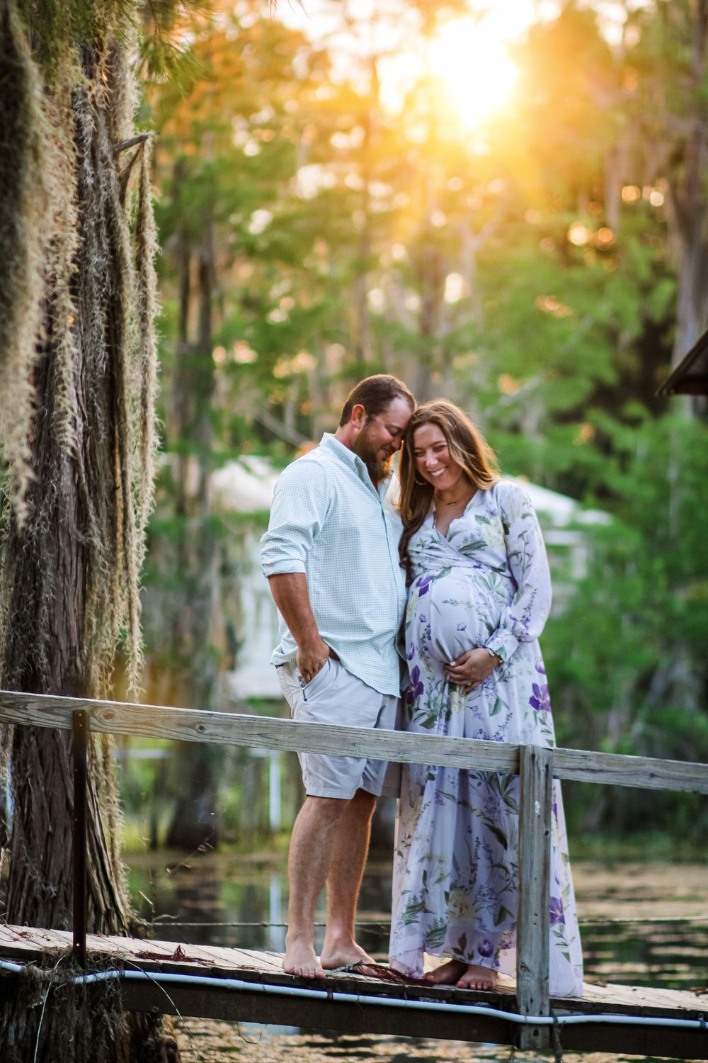 Expecting couple shares intimate moment on wooden dock during golden sunset in wooded area.