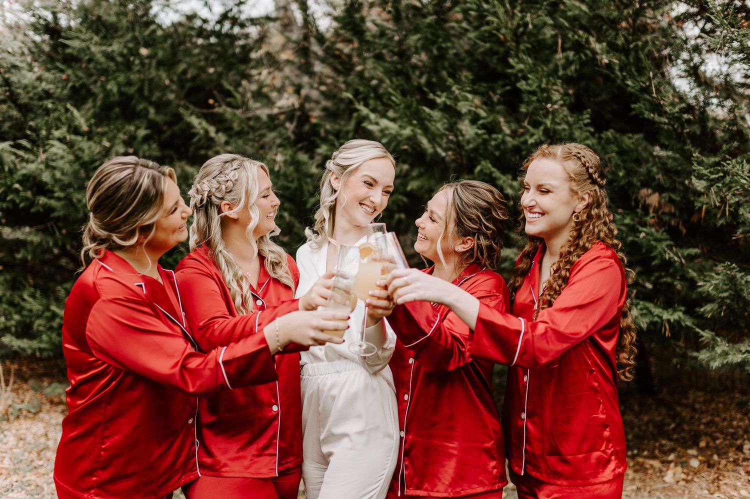 Wedding party in red and white robes celebrating outdoors among evergreen trees.