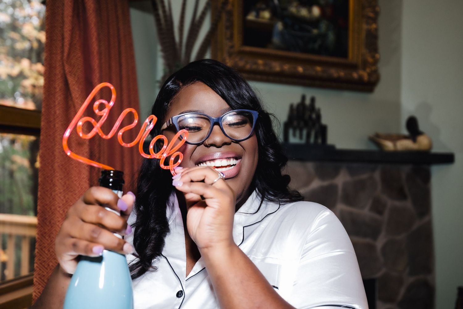 Joyful individual in glasses laughing while holding a neon sign in a bright living room setting.