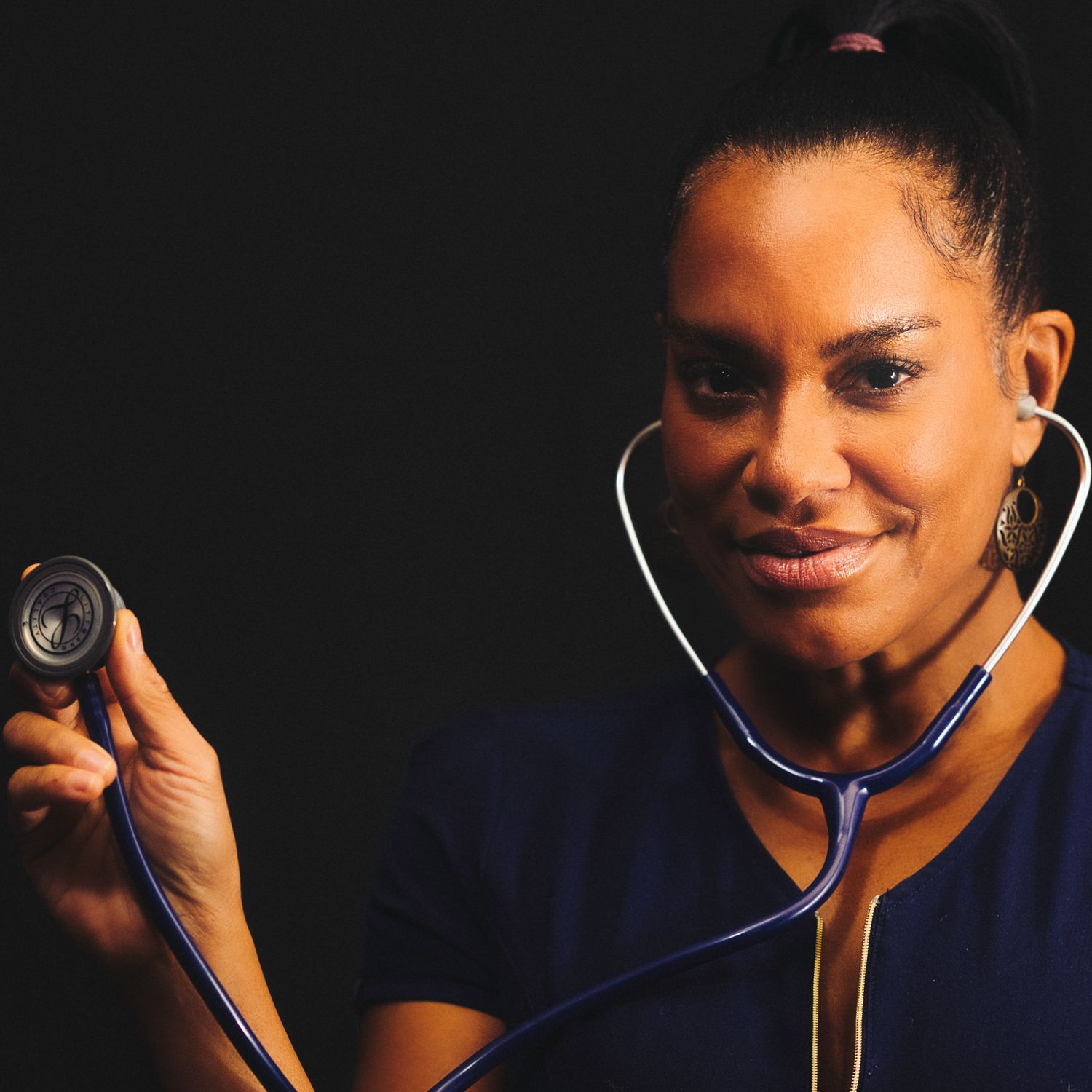 A healthcare worker in navy scrubs holds a stethoscope against a black background.
