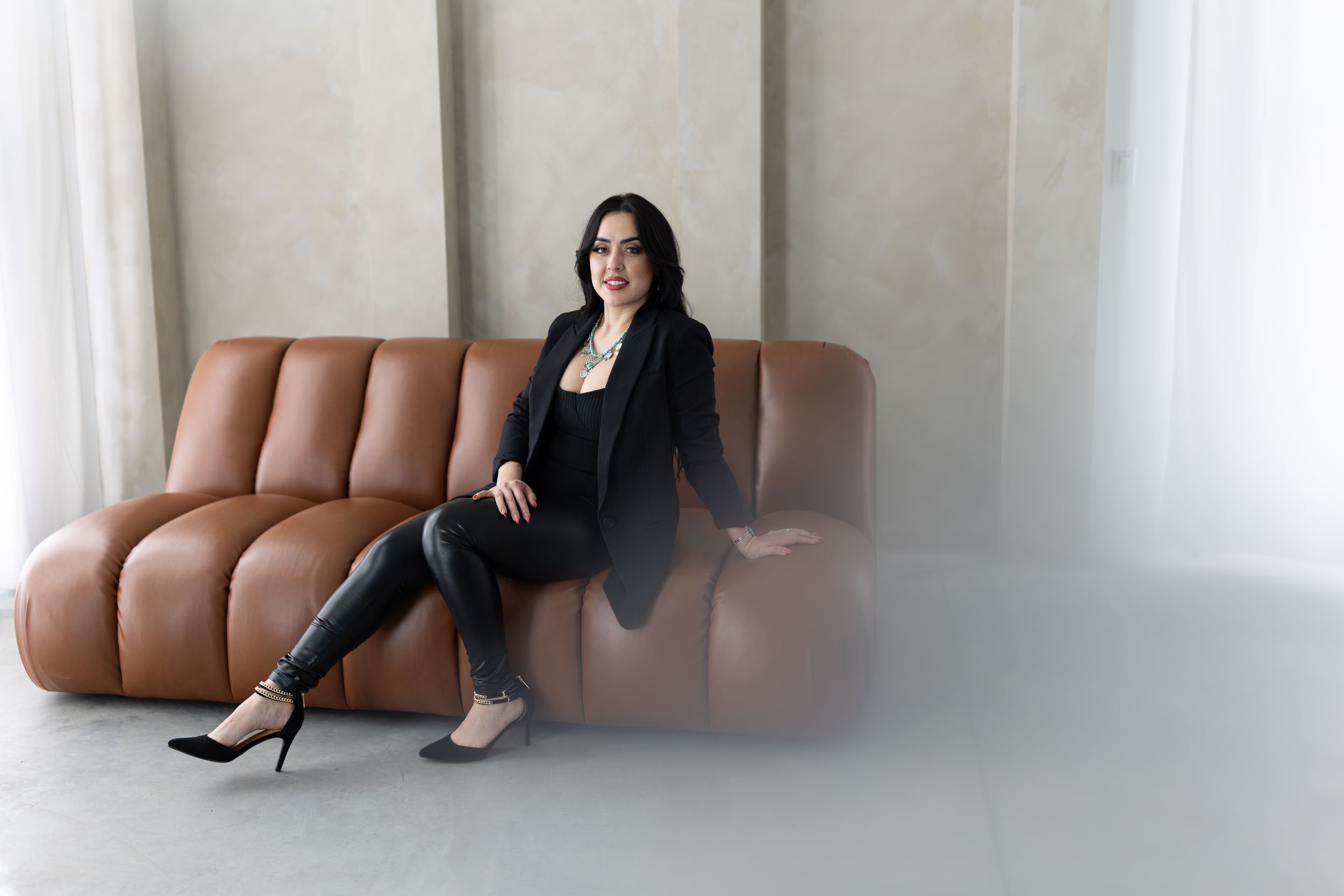 Professional figure seated on a tan leather couch wearing all black business attire in a minimalist setting.