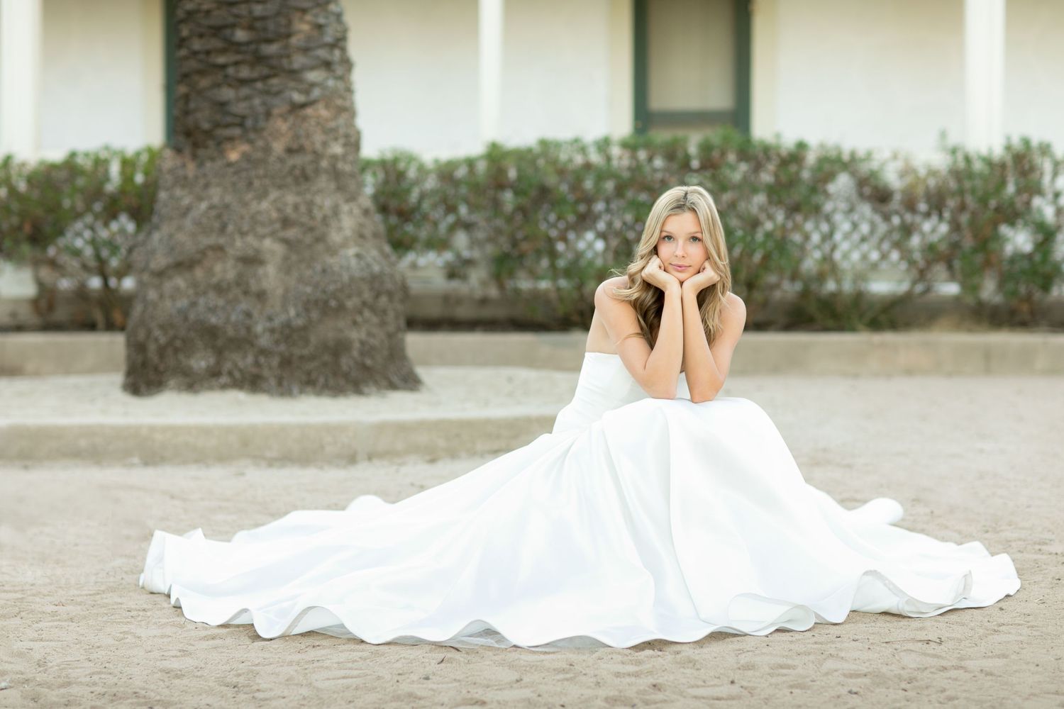 A person in a flowing white wedding gown sits gracefully on the ground with a palm tree in the background.