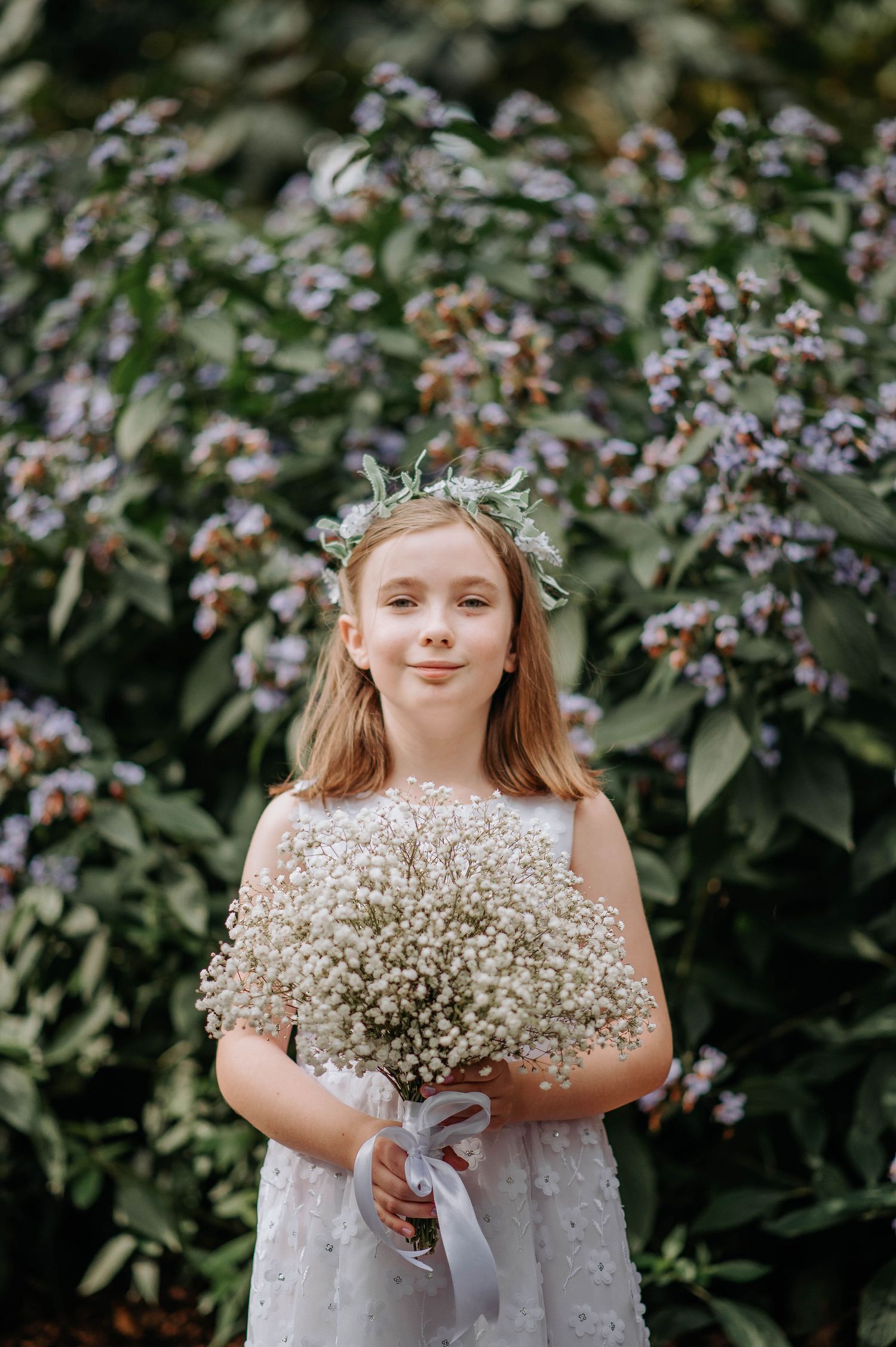 Young attendant in white dress holds baby's breath bouquet against flowering garden backdrop.