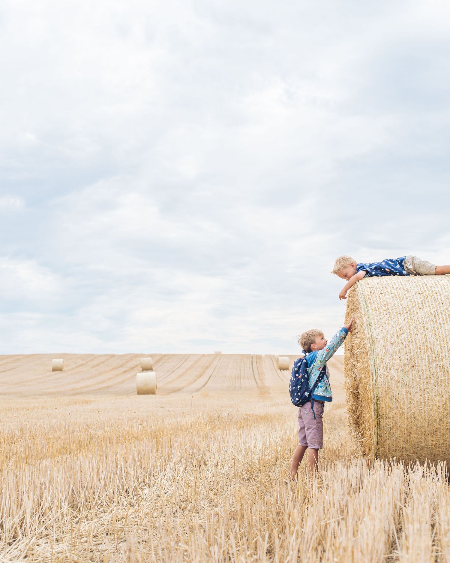 Two children play and climb on a large round hay bale in a golden wheat field under cloudy summer skies.