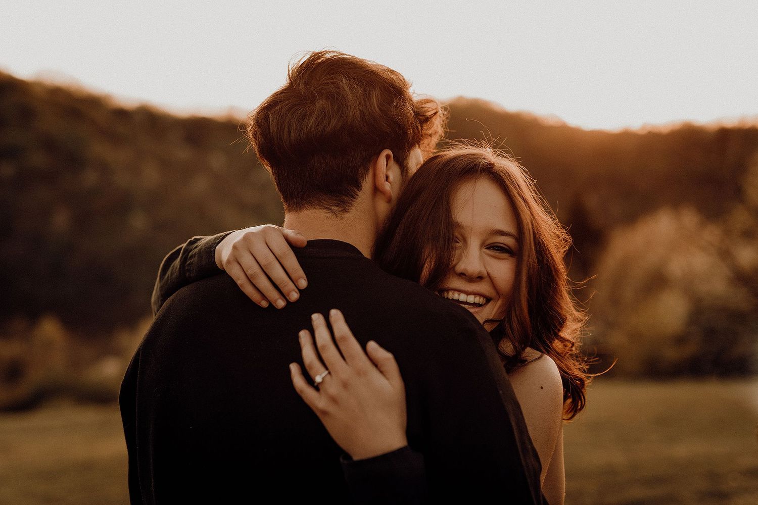 Happy couple embracing at golden sunset in outdoor field, woman smiling joyfully over mans shoulder.