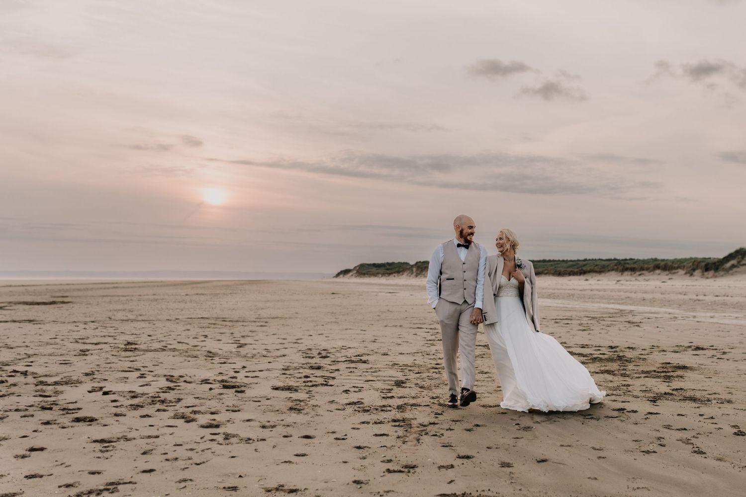 Wedding couple walking along sandy beach at sunset with ocean and cliffs in background.