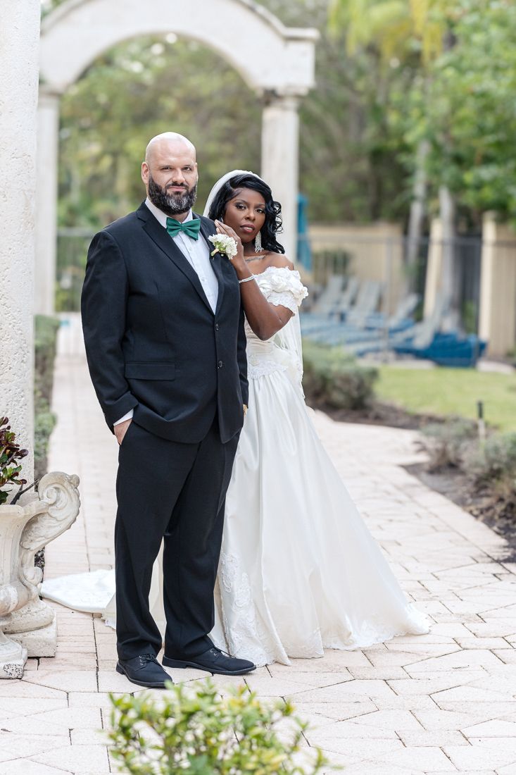 Bride and groom pose under archway in elegant wedding attire during outdoor ceremony.