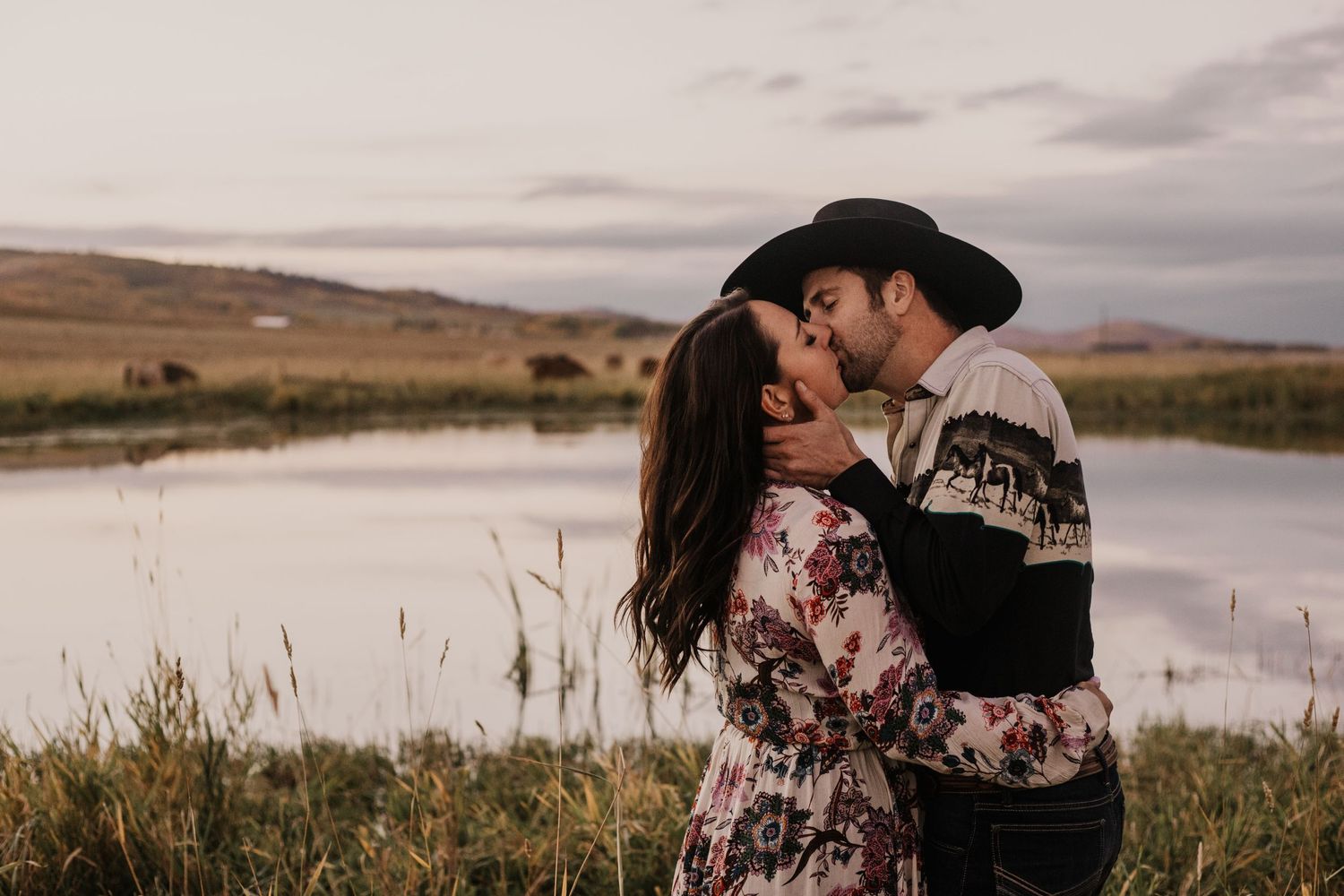 Cowboy kisses fiance in front of pond and field