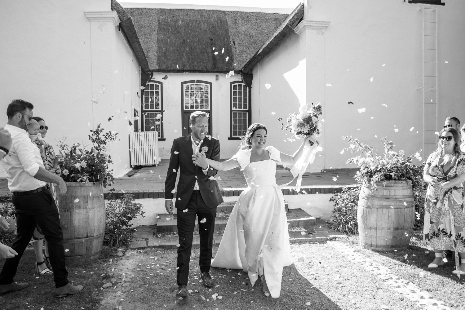Bride and groom joyfully walking through a shower of confetti in the sunlight.