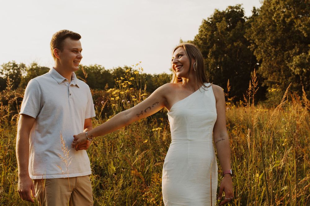 Couple holds hands in a golden field during sunset while wearing white and casual attire.