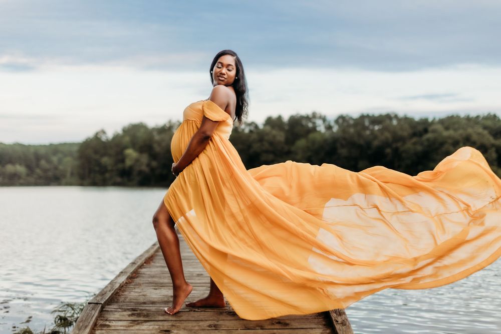 Flowing yellow dress catches wind as woman poses on wooden dock by lake.