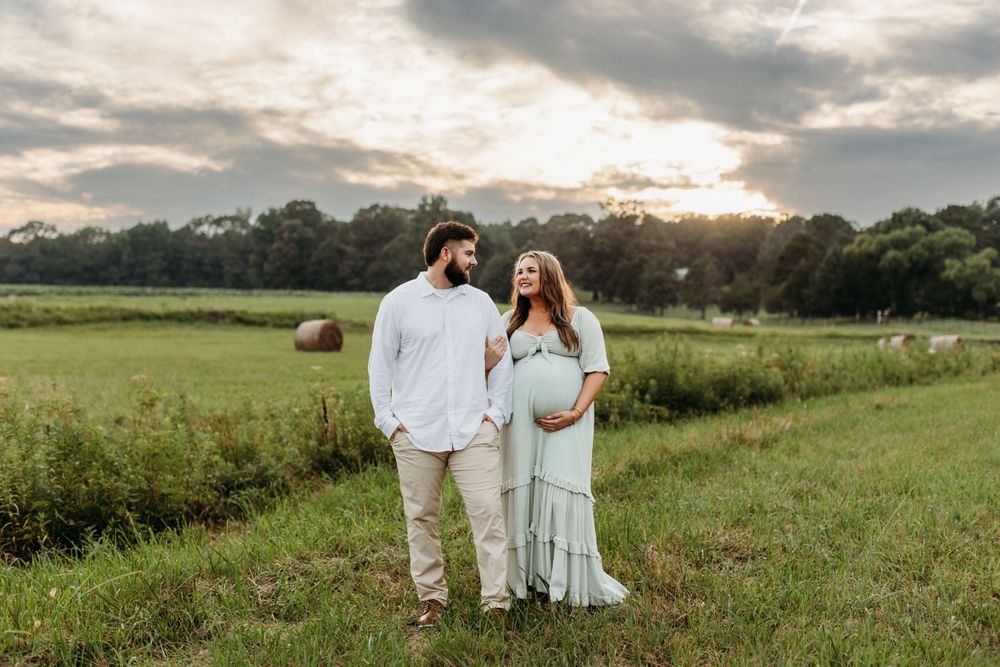 Sunset maternity photo with couple standing in green field with hay bales.