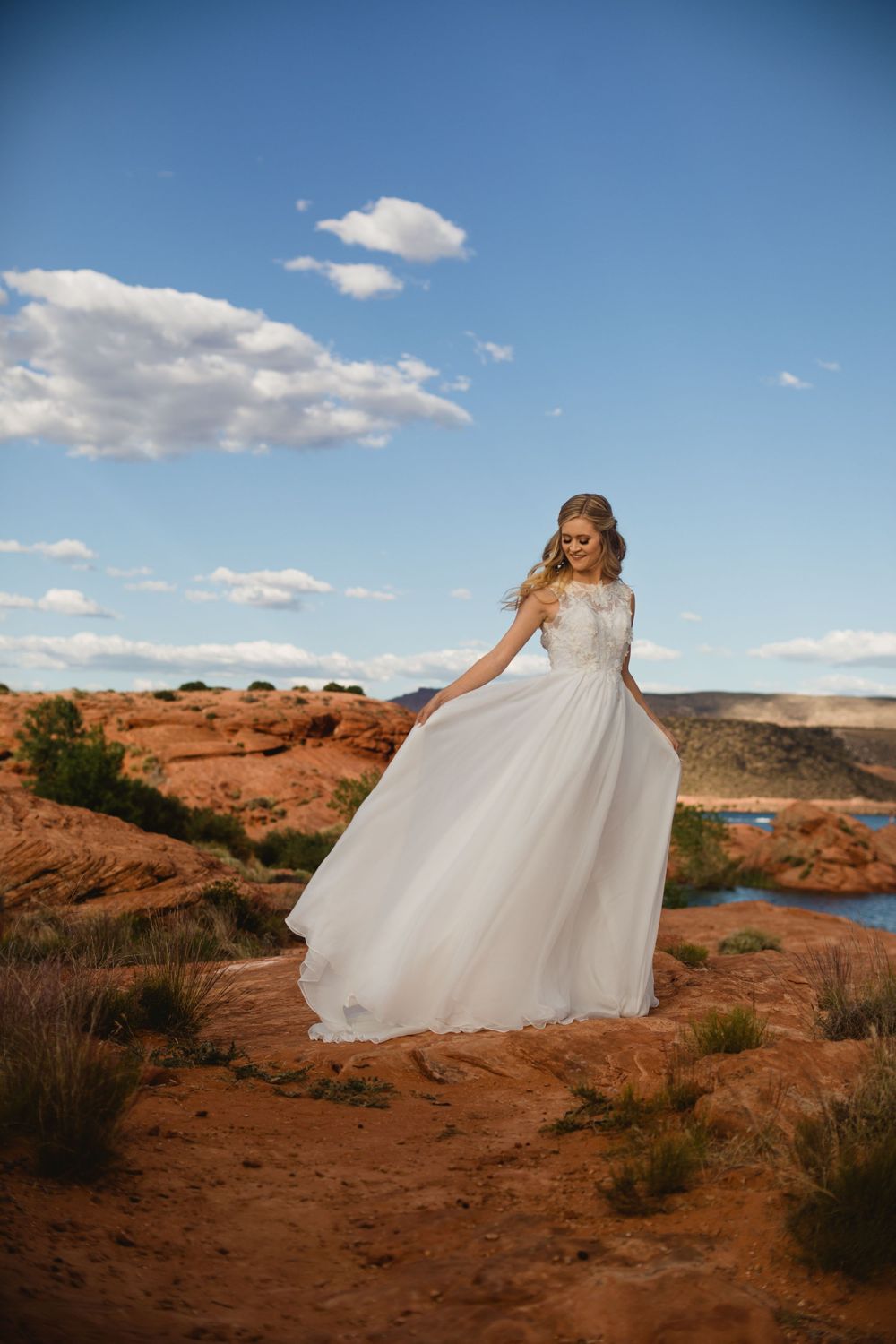 Bride in flowing white lace gown twirls on red rock desert landscape with blue sky and river.