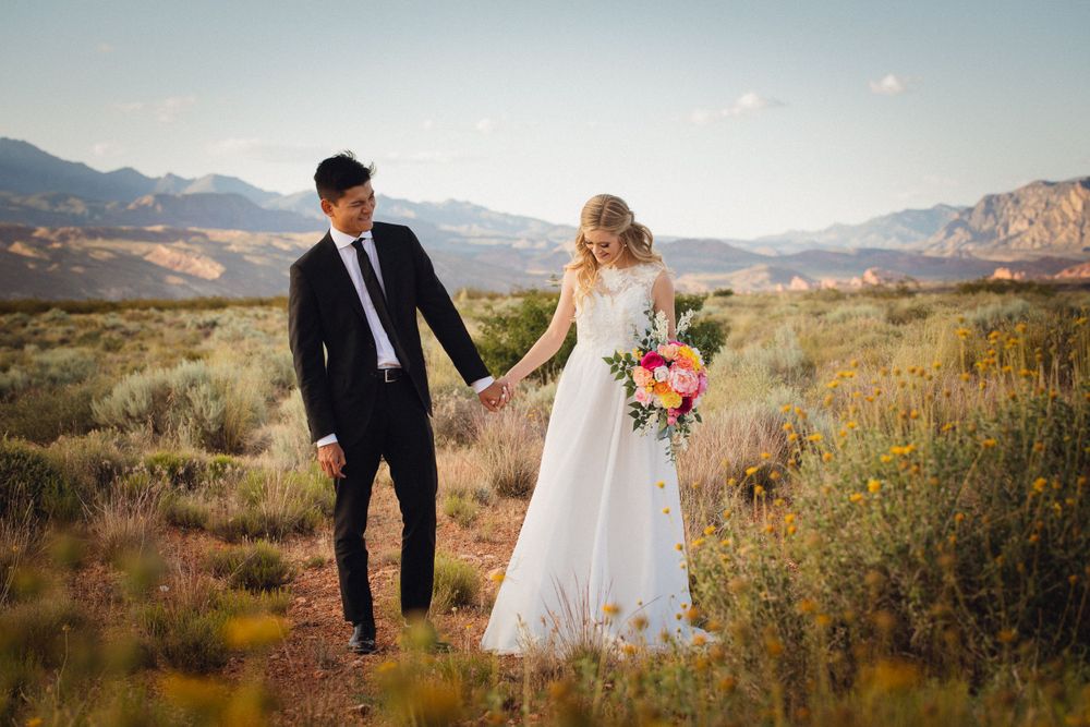 Wedding couple holds hands walking through desert brush with colorful mountain scenery behind them.