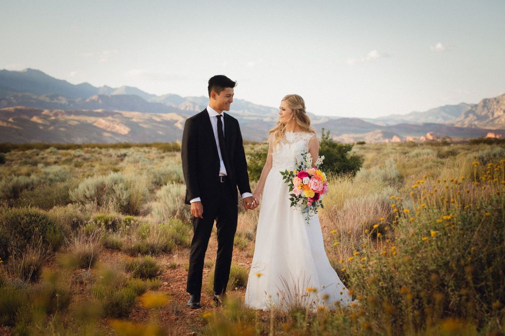 Bride holding vibrant bouquet and groom hold hands gazing at each other in a scenic desert setting.