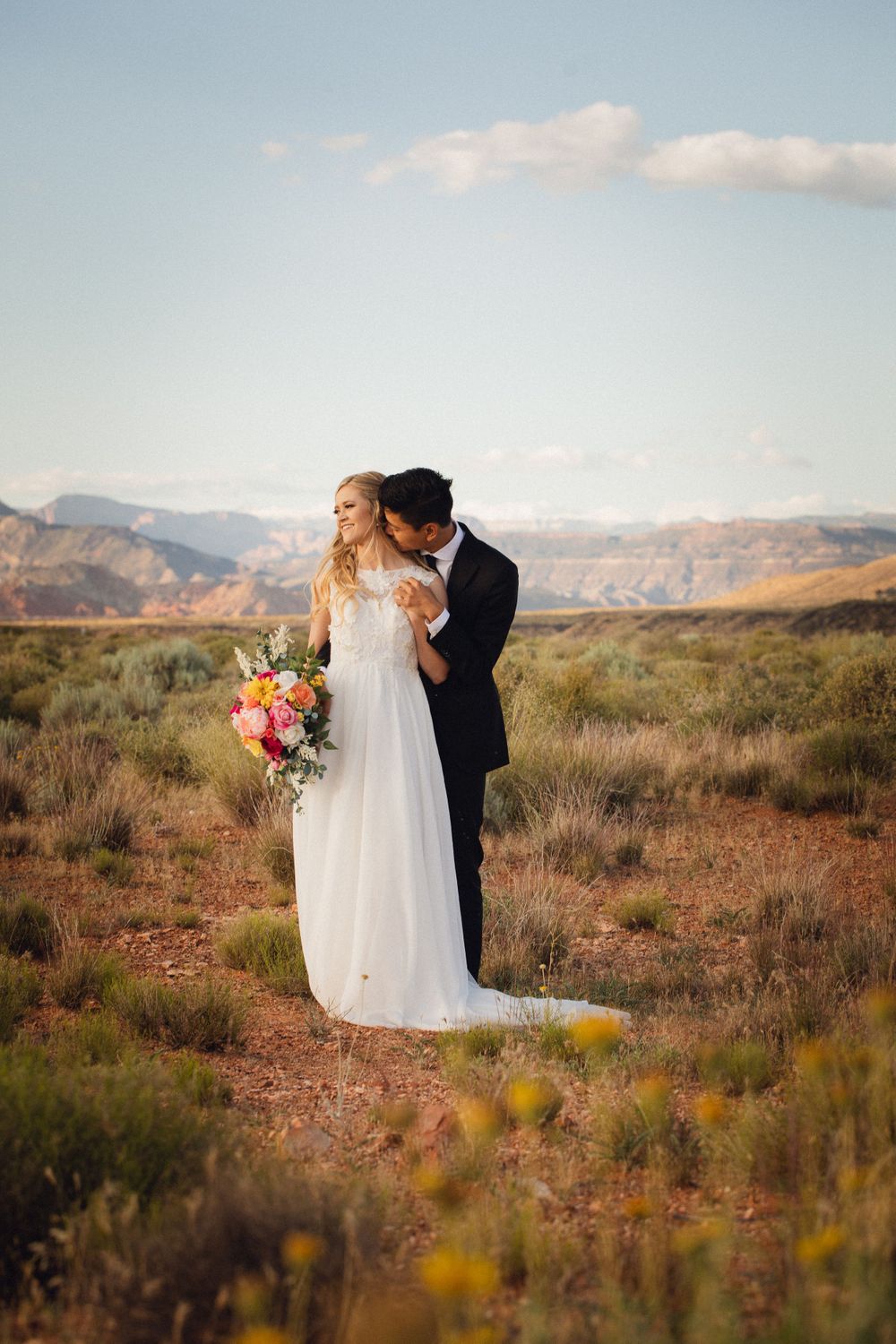 Groom embraces bride from behind in open desert landscape with mountains and blue sky above.