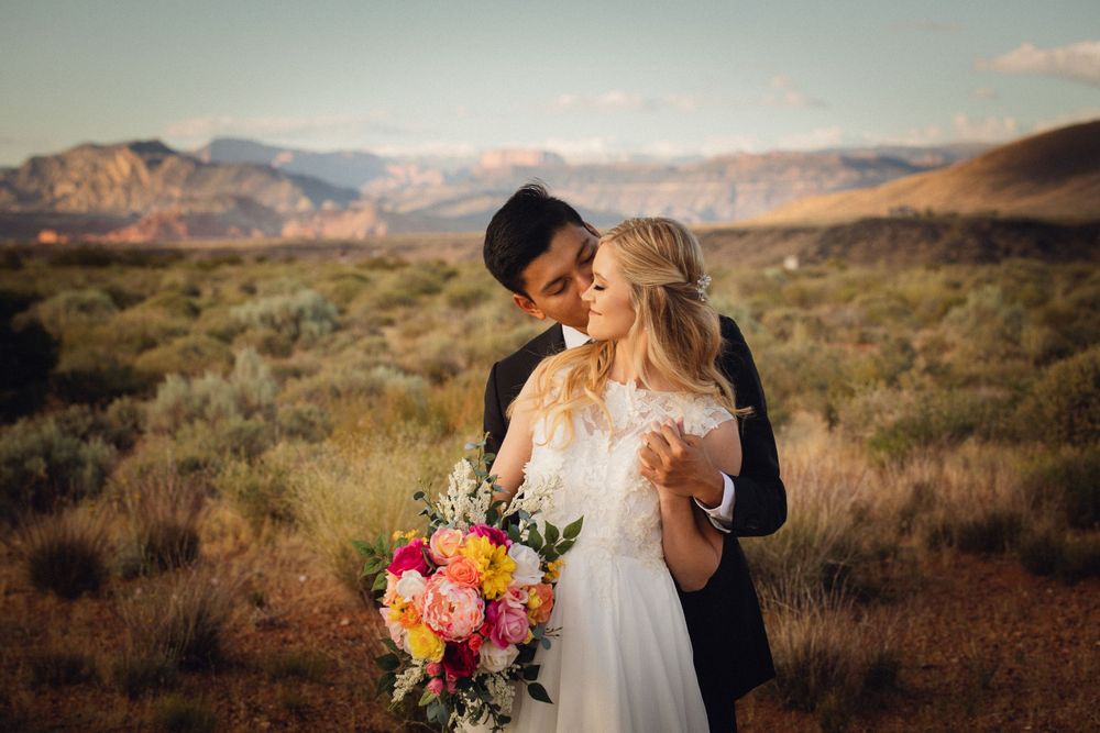 Groom tenderly kisses bride holding bright floral bouquet with red rock mountains in the background.