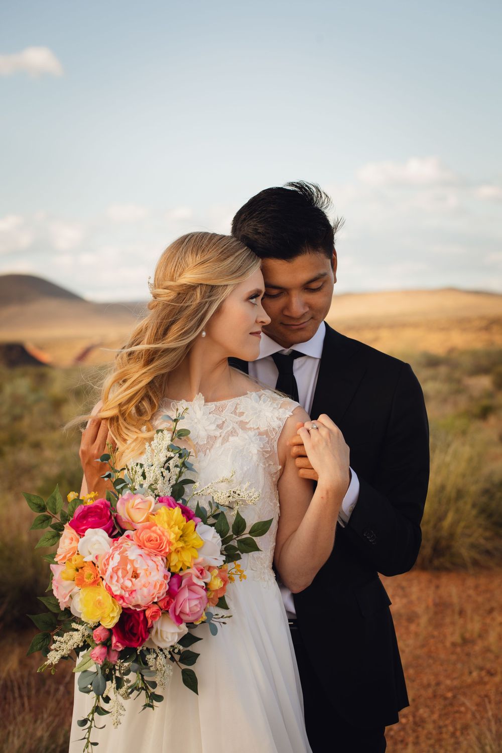 Bride and groom share a close romantic moment with a vibrant pink and yellow bouquet in the desert.