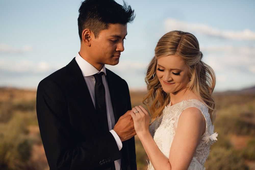 Groom in black suit examines bride's ring while couple shares intimate outdoor desert moment.