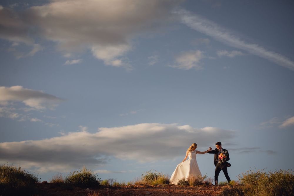 Bride and groom dance hand in hand on sandy desert path under blue sky.