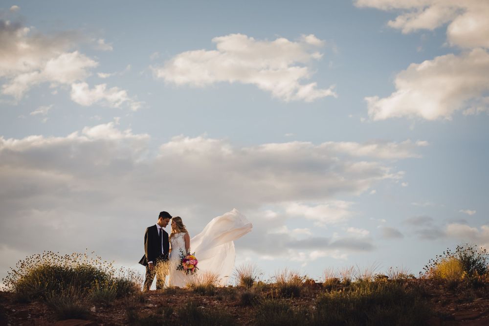 Bride and groom kiss on desert hilltop with flowing veil and blue cloudy sky backdrop.