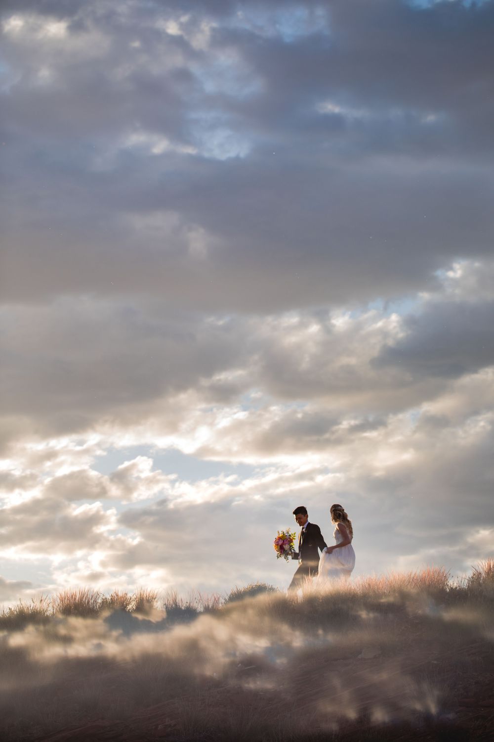 Silhouetted bride and groom stand together on hilltop beneath dramatic cloudy sky at dusk.