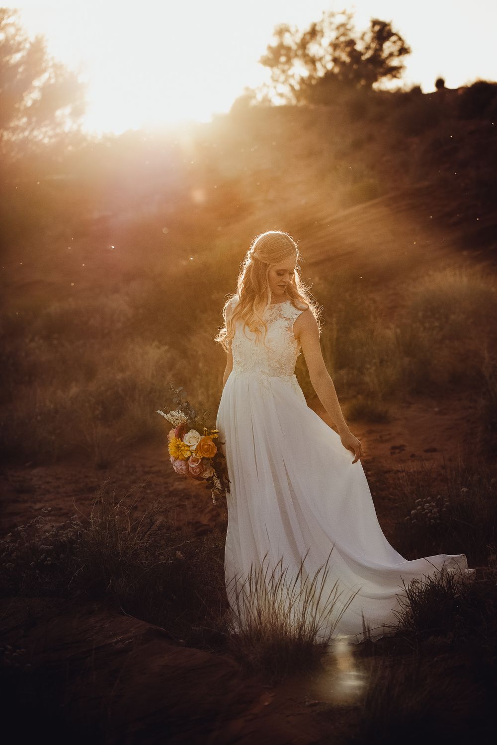 Bride in lace gown holds bouquet bathed in golden sunset light rays in desert.