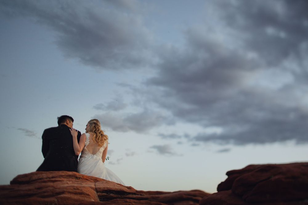 Bride and groom sit together on red sandstone rocks beneath dramatic stormy twilight sky at dusk.