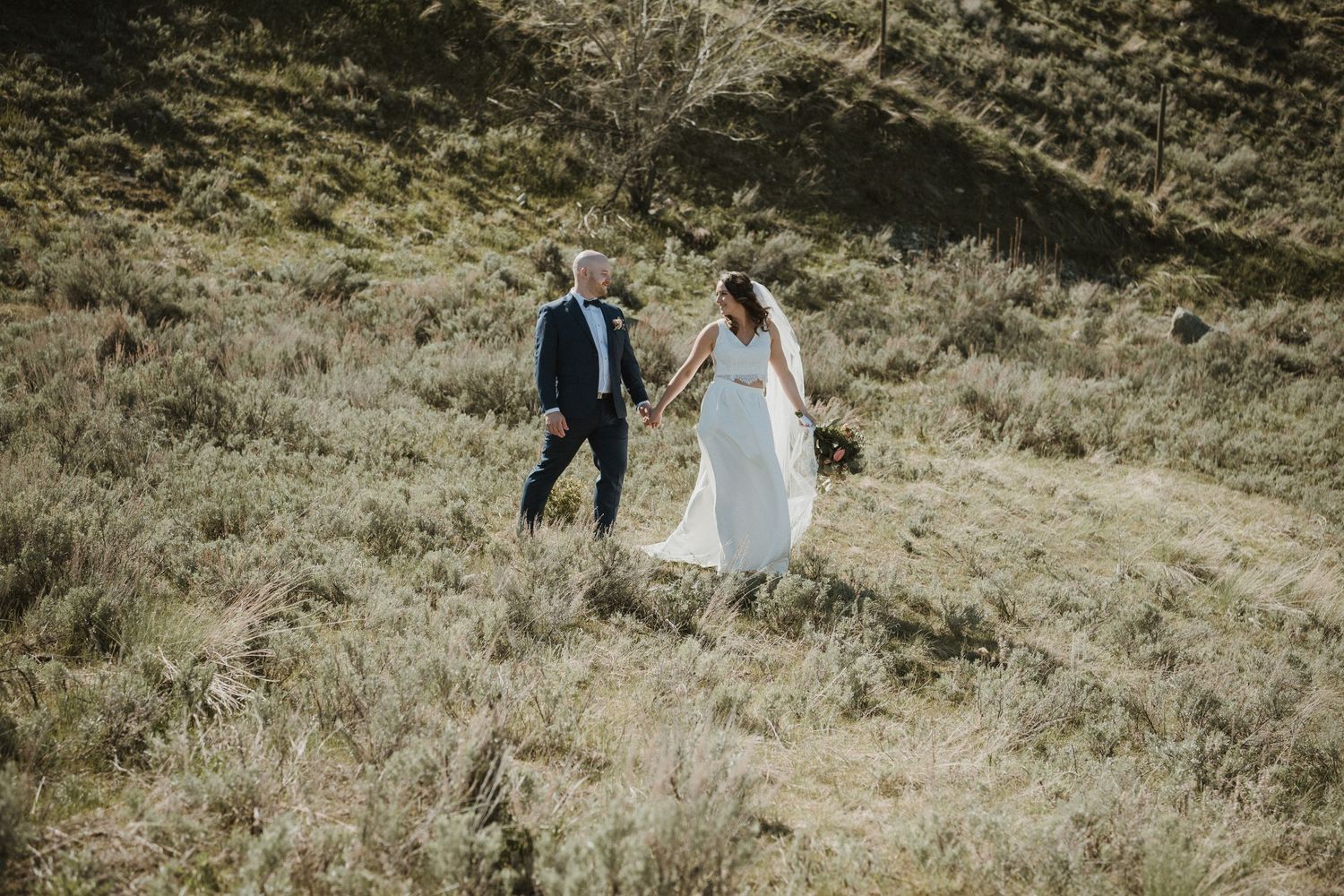 Newlywed couple walking together on a grassy hillside in formal wedding attire.
