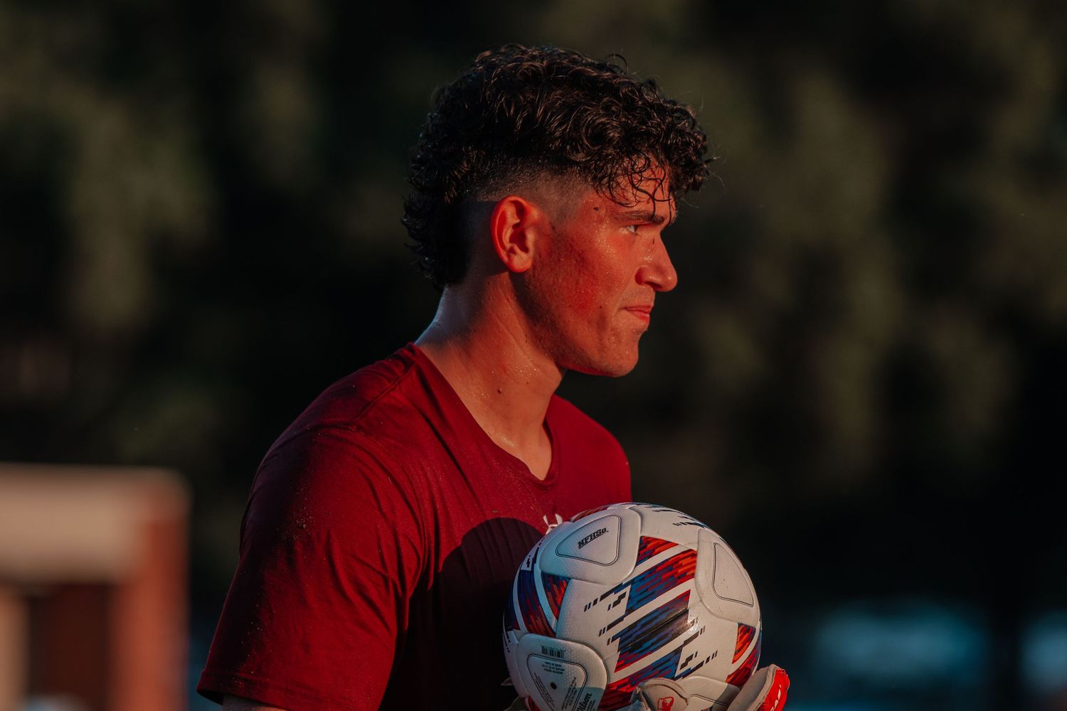 Soccer player in red jersey holds a soccer ball during golden hour lighting.