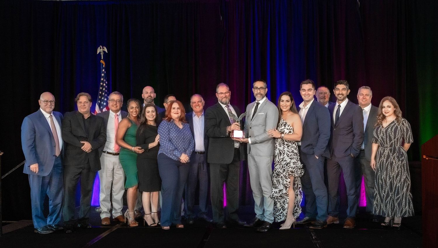 Group of professionals in formal attire pose on stage holding a trophy award at a gala event.