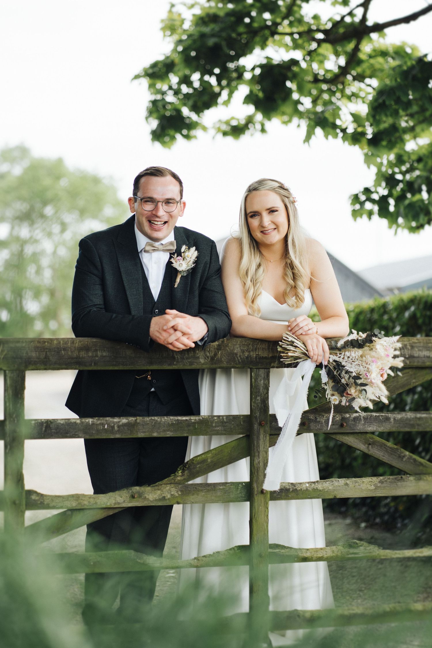 Newlywed couple posing together at rustic wooden fence during outdoor wedding.