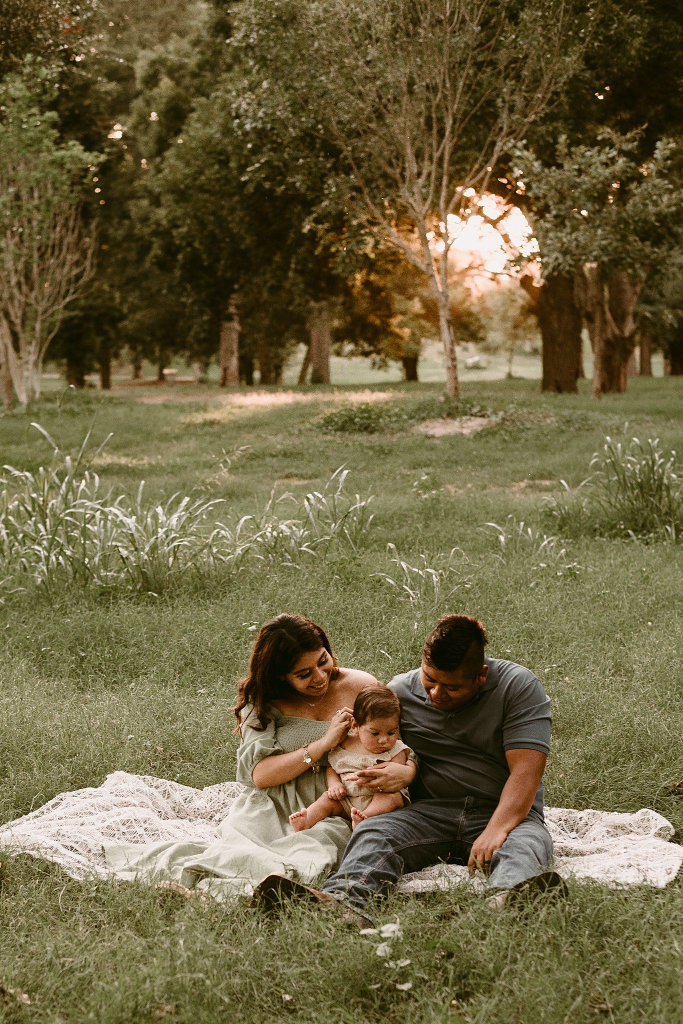 Family relaxing on white blanket in park with golden sunset filtering through trees.