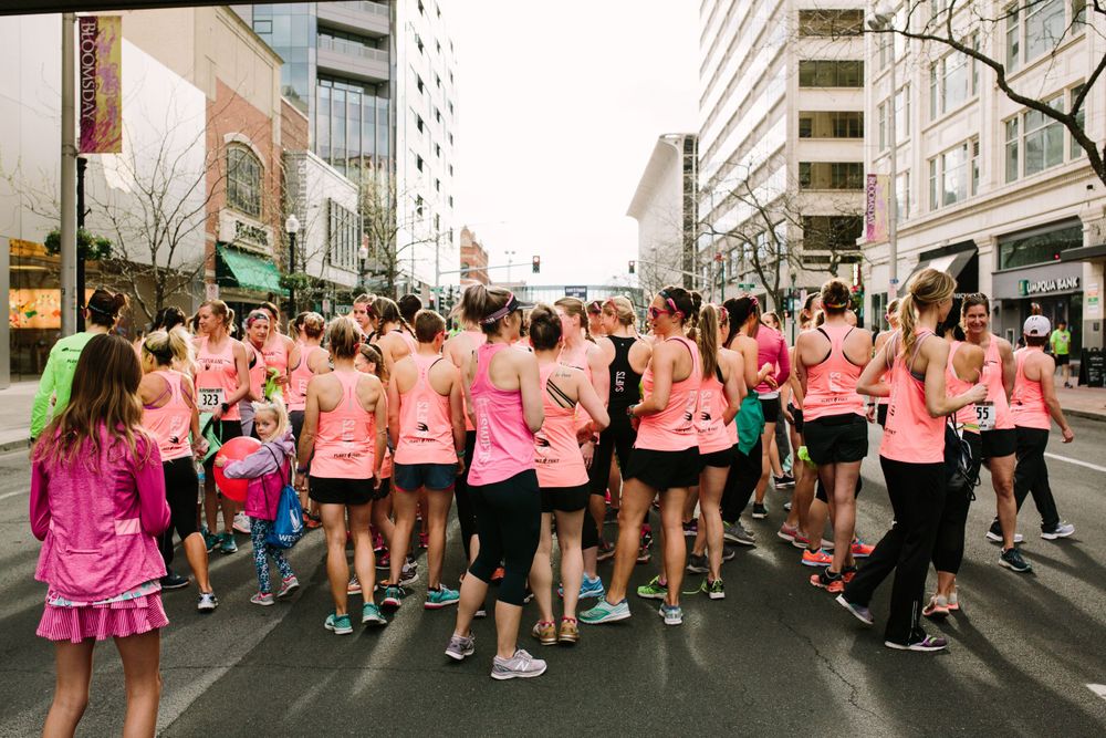 Large group of runners in pink tops walking along city street during organized race event.