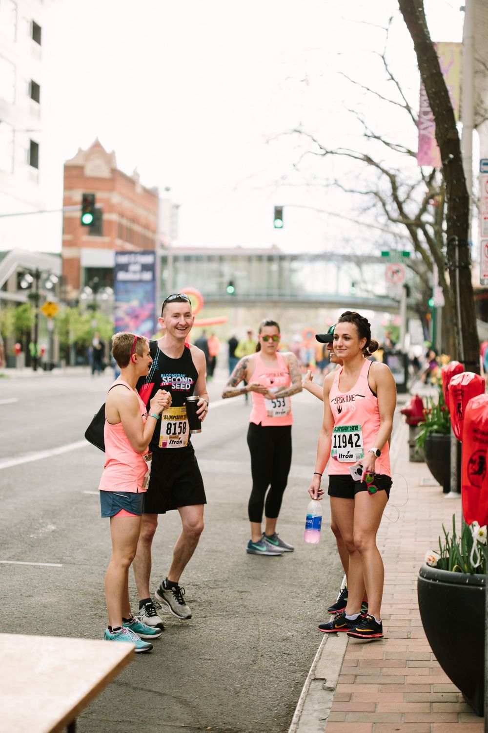 Runners in pink and black race bibs socializing on city sidewalk during street running event.
