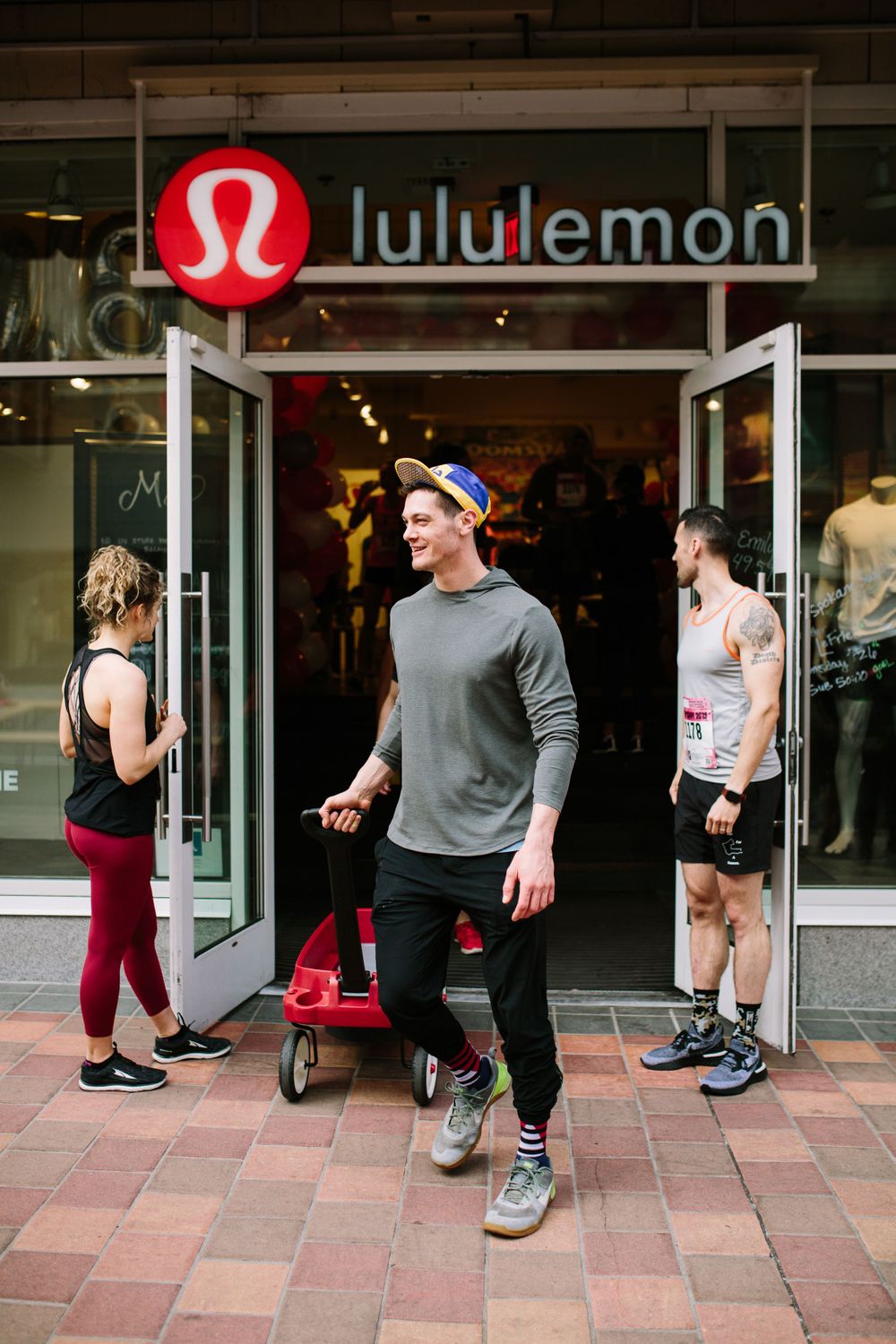 Man pulling red wagon exits Lululemon store while two others stand near open glass doors on brick sidewalk.
