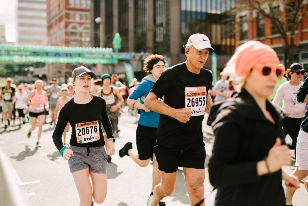 Runners wearing race bibs compete in a city marathon on a sunny street with green bridge in background.