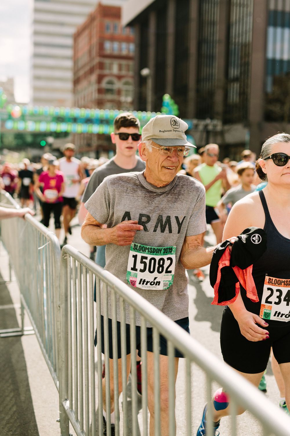 Elderly man in Army shirt with race bib 15082 runs Bloomsday race alongside other participants near metal barriers.
