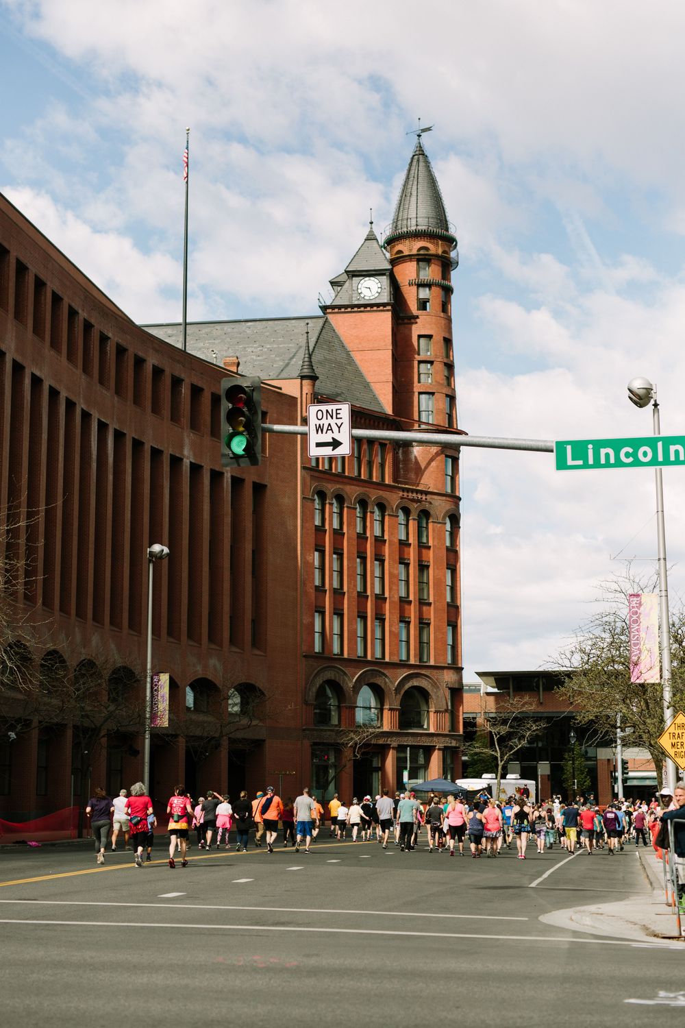 Runners cross intersection at Lincoln Street past historic red brick clock tower building on cloudy day.