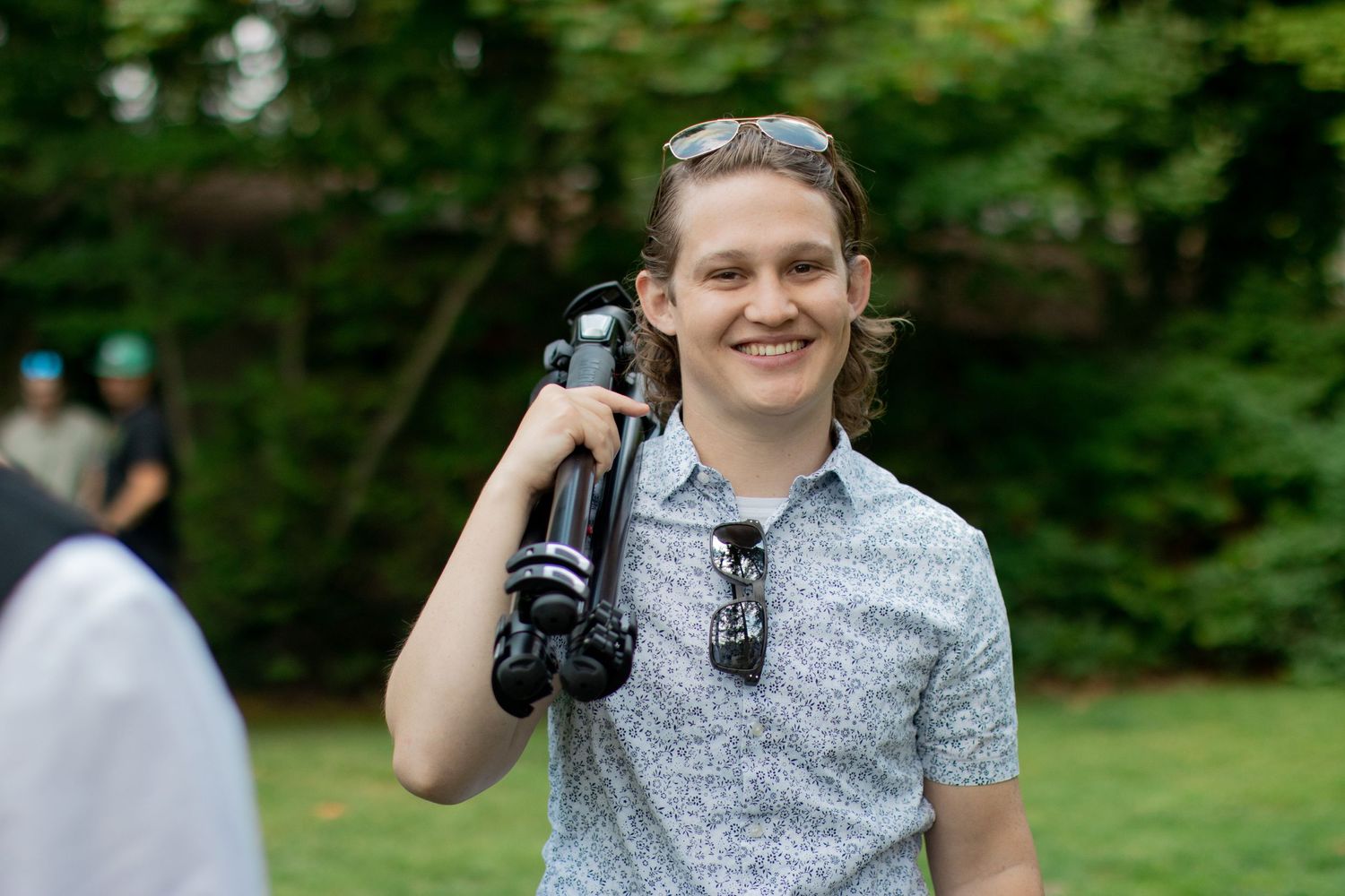 Young man smiling outdoors carrying a camera tripod over his shoulder at a garden event.