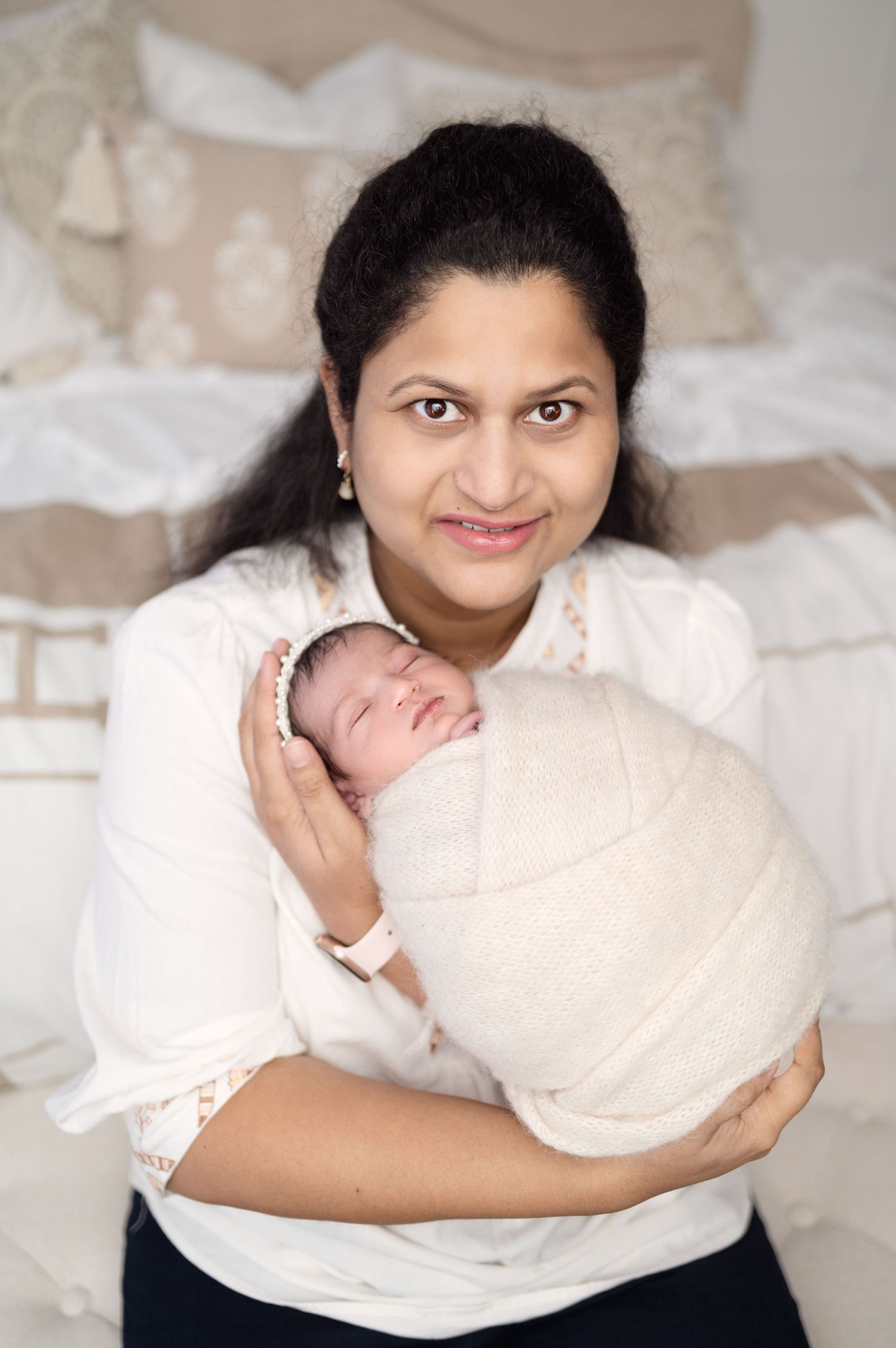 Parent in white blouse smiles while cradling sleeping newborn wrapped in cream blanket.
