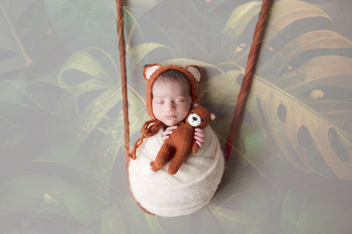 Baby in white outfit lying in hanging basket with teddy bear amid tropical leaves.