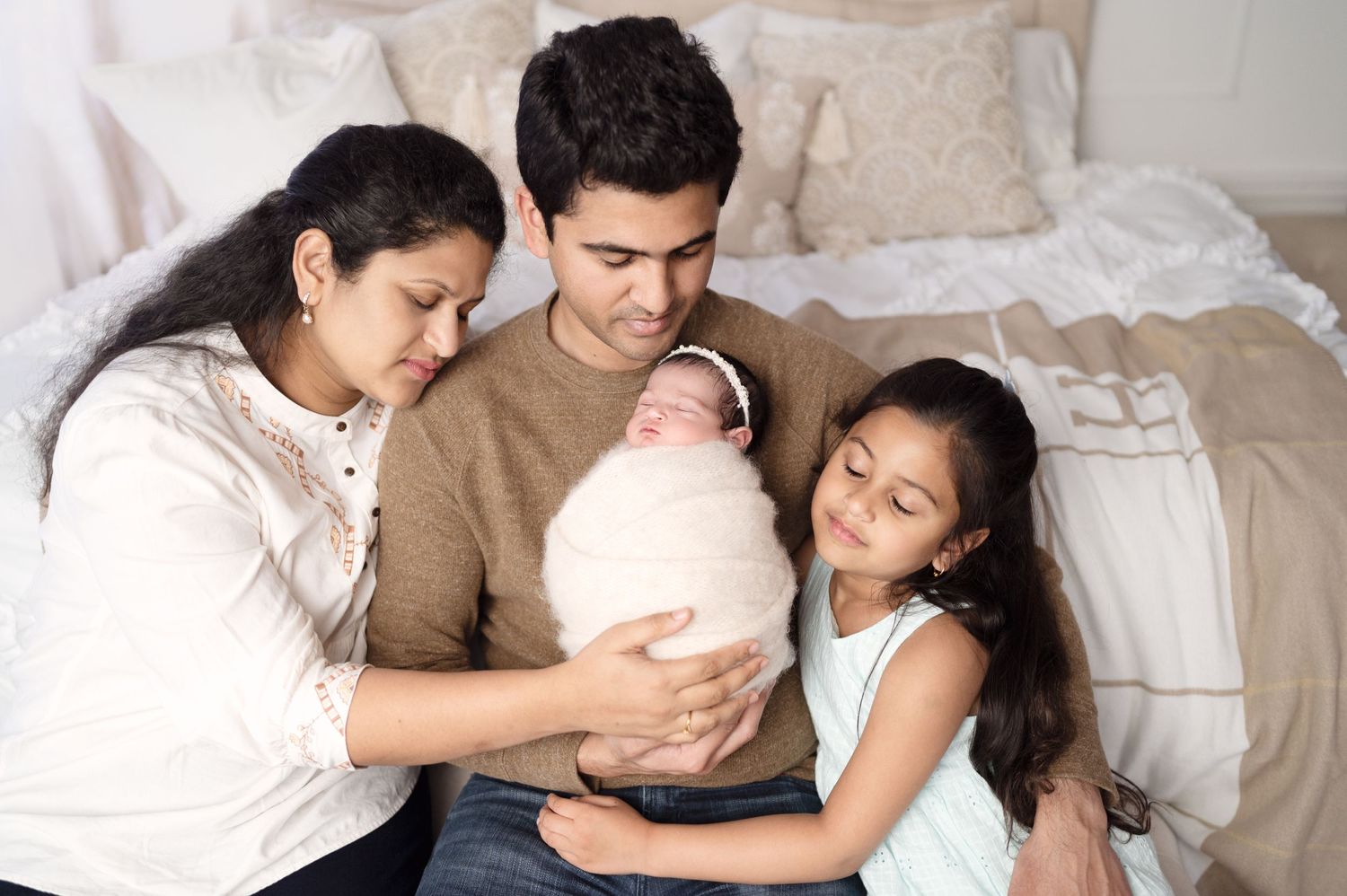 Family gathers on bed to admire their newborn baby wrapped in white.