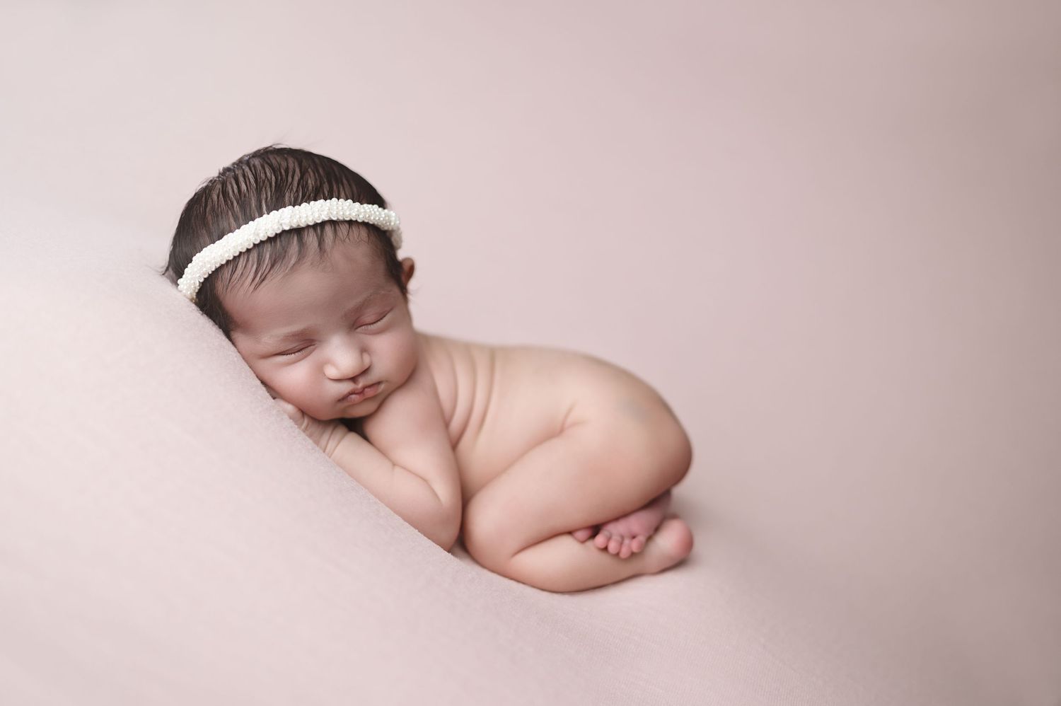 Peaceful newborn sleeping on side with white headband against pink backdrop.