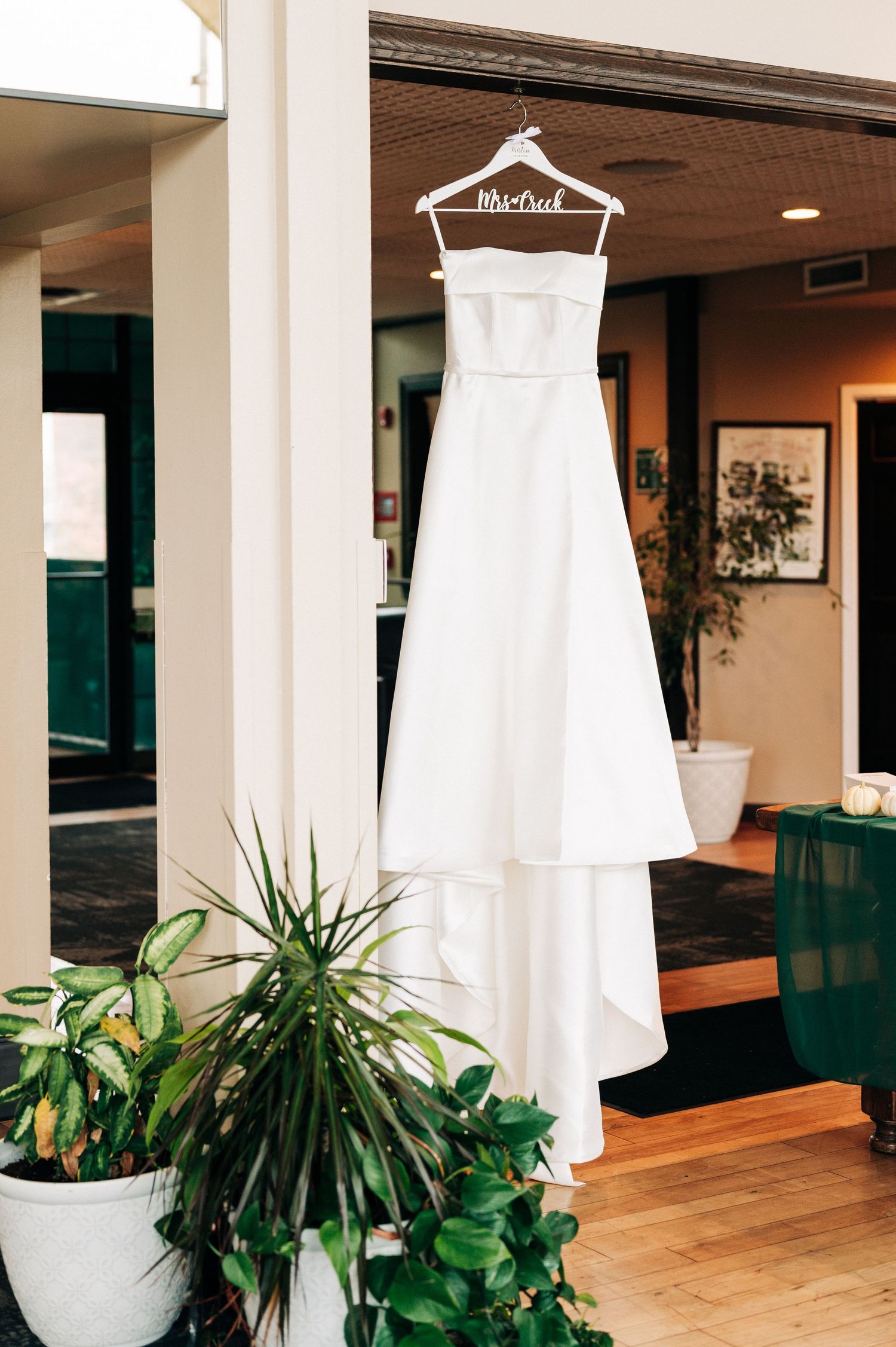 An elegant white wedding dress hangs on display with greenery and natural lighting in the background.