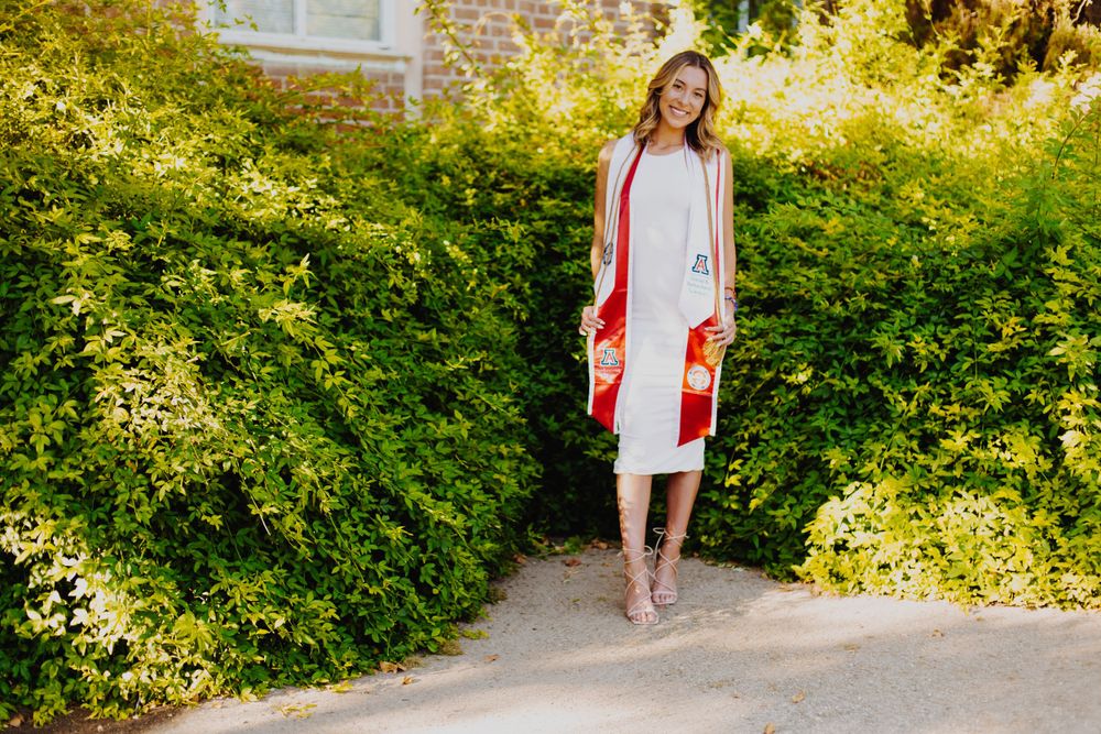 A woman in a white dress with bold red stripes stands on a garden path surrounded by lush greenery.