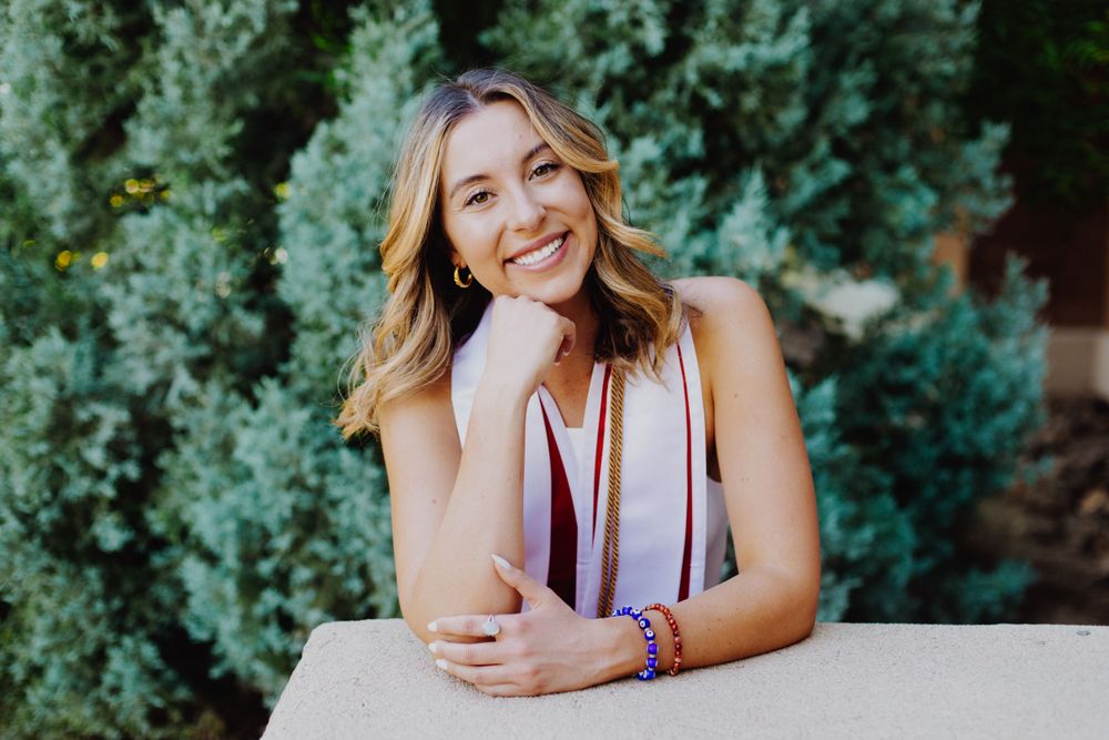 A casually dressed person poses with a bright smile against a backdrop of evergreen shrubs.