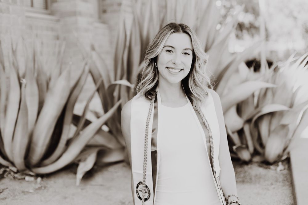 Black and white graduation portrait with academic regalia against natural backdrop.