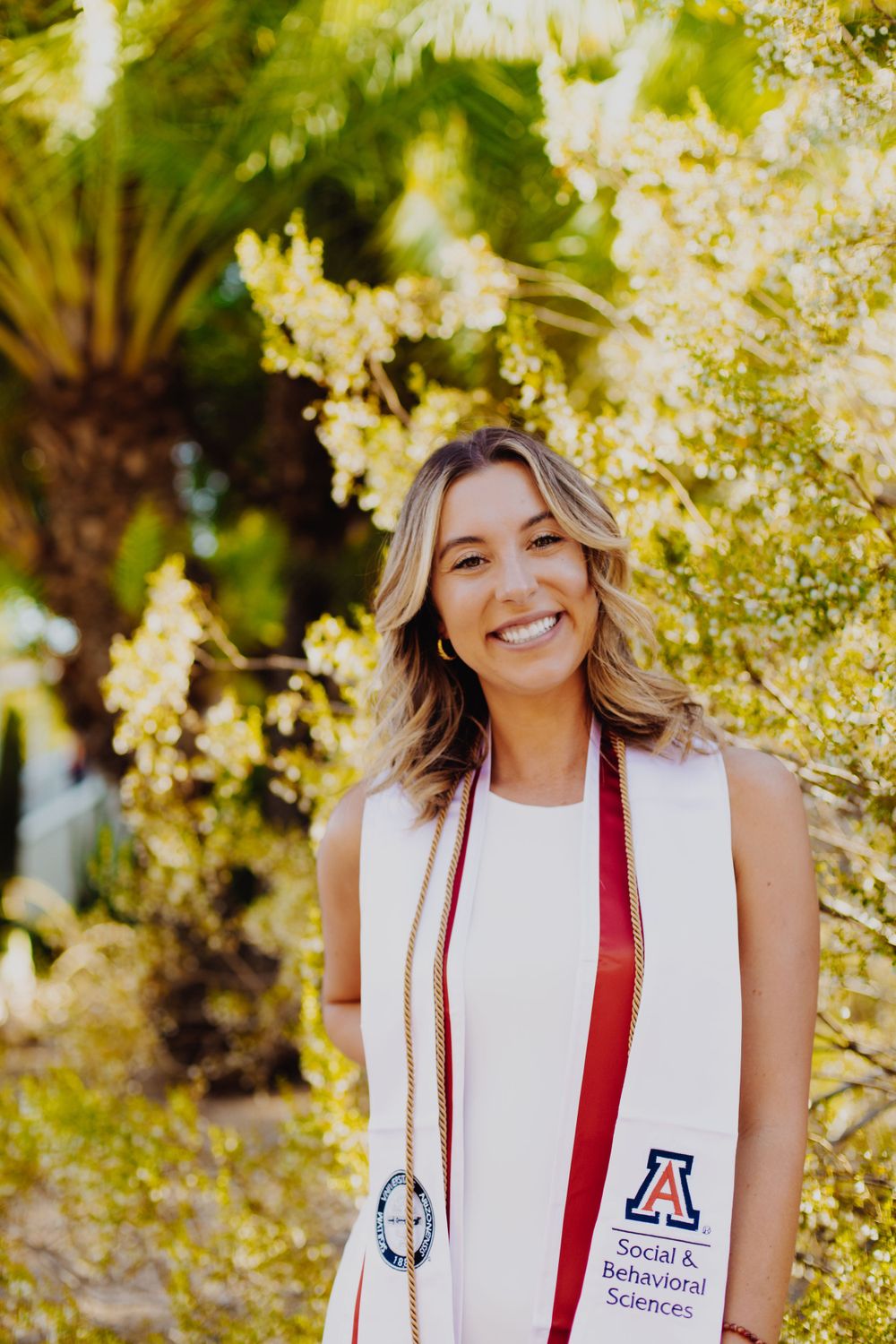 Graduate wearing University of Arizona stole poses among sunlit trees.
