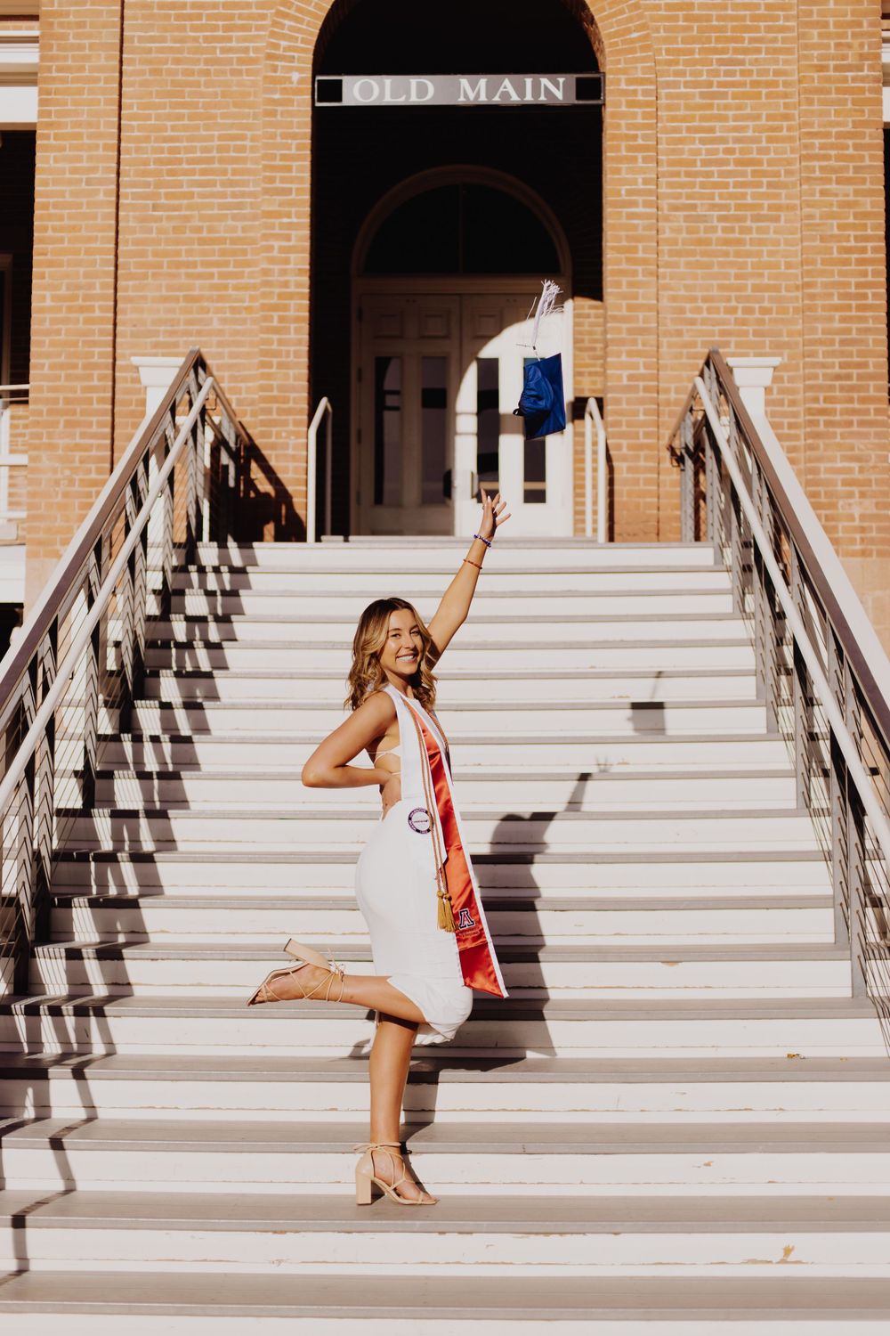 A person strikes a celebratory pose on the steps of Old Main building wearing a white dress with red accents.
